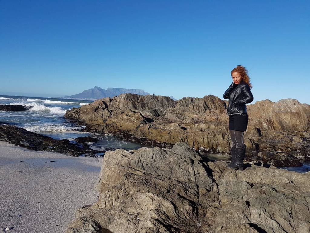 A person standing on rocky beach terrain with ocean waves crashing in the background, and Table Mountain visible in the distance under clear blue skies.