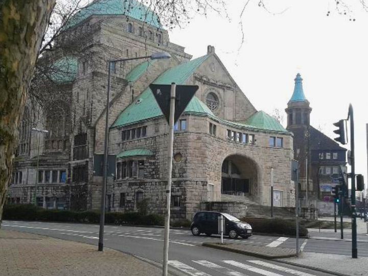 Exterior view of a historical building with a green roof, featuring large stone walls and an arched entrance, surrounded by street signs and a parked car.