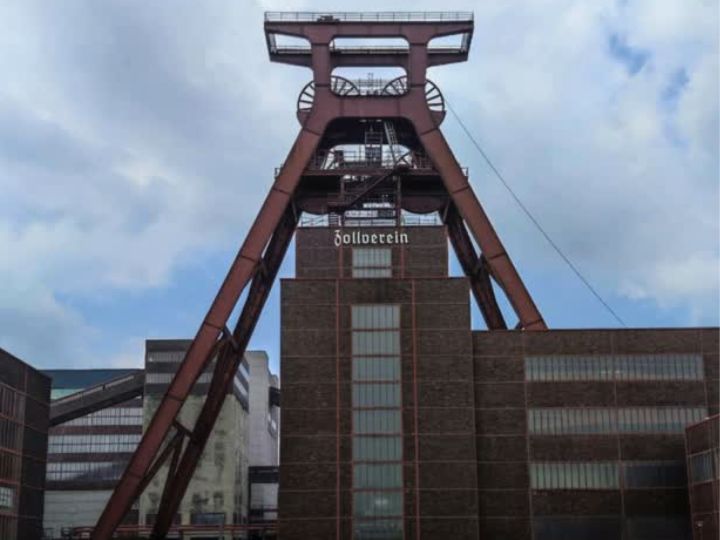 Exterior view of the Zollverein Coal Mine in Essen, showcasing its iconic red industrial structures against a cloudy sky.