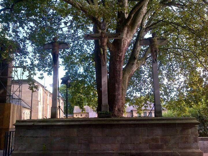 A large tree with a mix of greenery and three crosses in front, situated on a stone base, with a building visible in the background.