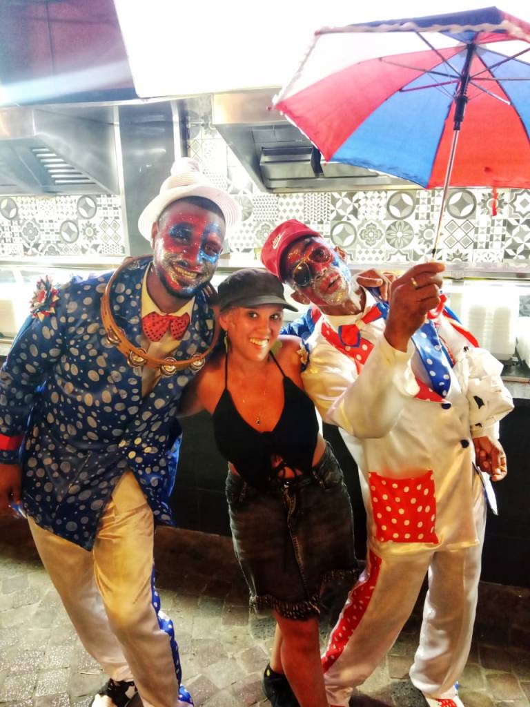 A group of three individuals, two men in colorful, polka-dotted costumes with face paint, and a woman in casual clothing, smiling and posing together in a festive atmosphere.