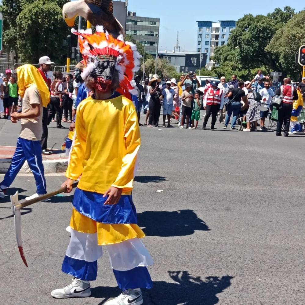 A performer dressed in a bright yellow and blue costume with a colorful feathered headpiece, holding a traditional tool, stands in the middle of a street during the Tweede Nuwe Jaar celebration in Cape Town. A crowd can be seen in the background, enjoying the vibrant festivities.
