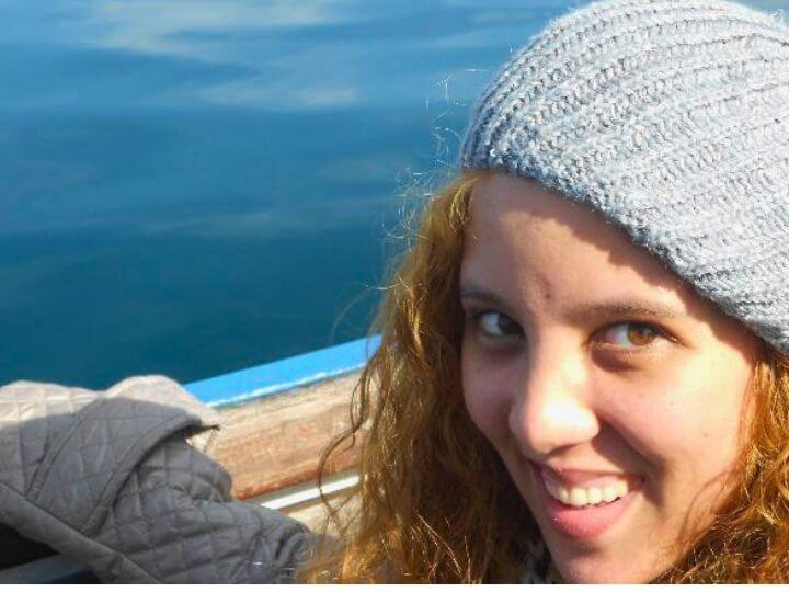 A smiling person with curly hair wearing a knitted hat, sitting on a boat with calm blue water in the background.