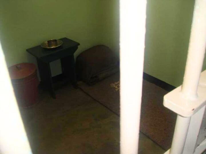 Interior of Nelson Mandela's prison cell on Robben Island, featuring a small bed with a blanket, a basic table with a bowl, and a bucket, viewed through the bars.