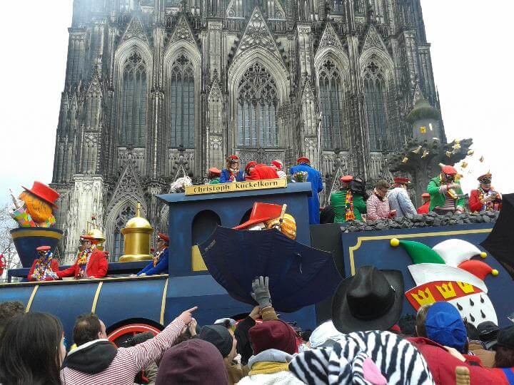 A colorful float with costumed performers during the Cologne Carnival, in front of the Cologne Cathedral, with a large crowd of festival-goers enjoying the festivities.