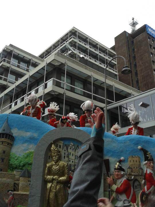 Participants in traditional costumes on a festive float during Cologne Carnival, with a city backdrop.