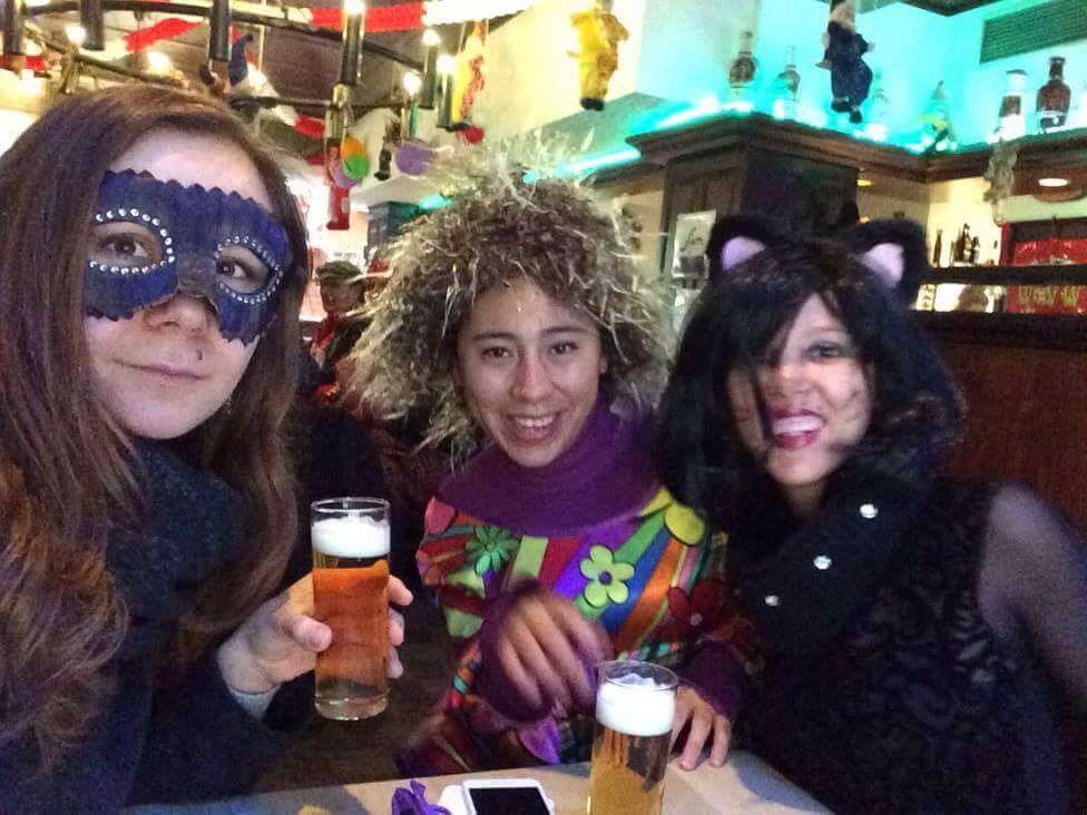 Three friends celebrating Cologne Carnival with drinks, wearing colorful costumes and masks in a festive setting.