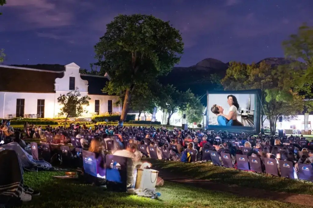 Outdoor cinema experience with a large movie screen set up at night, surrounded by a crowd of people sitting on folding chairs in a park setting.